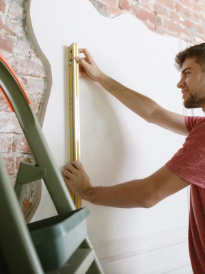 Young man preparing for doing apartment repair by hisselfes. Before home makeover or renovation. Concept of relations, family, DIY. Measuring the wall before painting or design making.